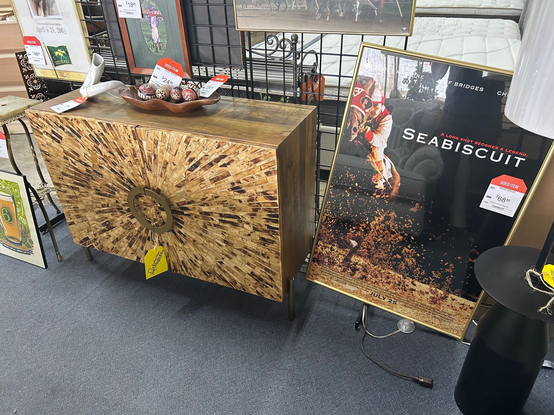 A modern wooden cabinet with a circular design pattern sits next to a framed Seabiscuit movie poster in a shop.