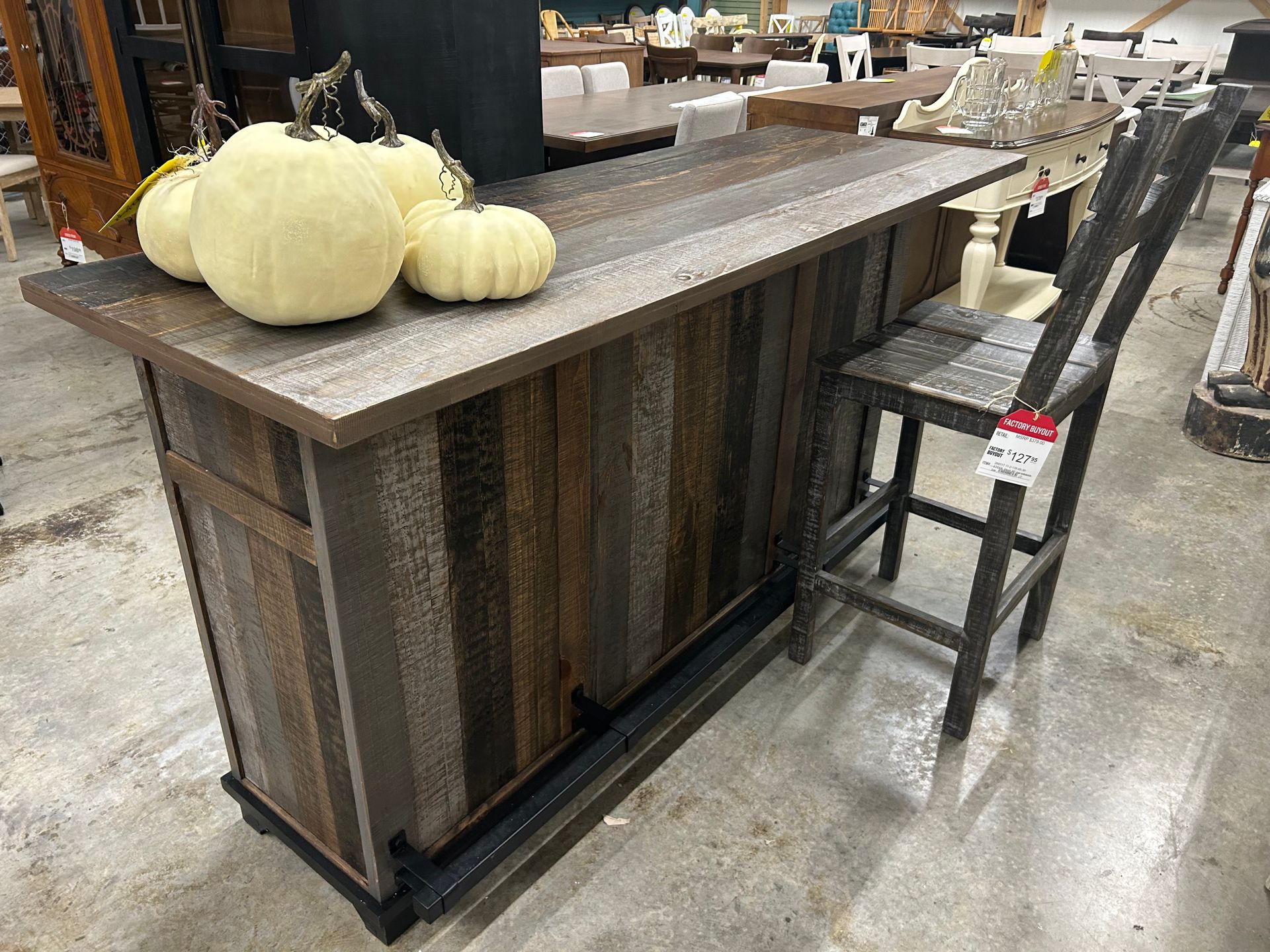 A rustic bar made of reclaimed wood and corrugated metal, topped with three white pumpkins, stands with a tall bar stool.