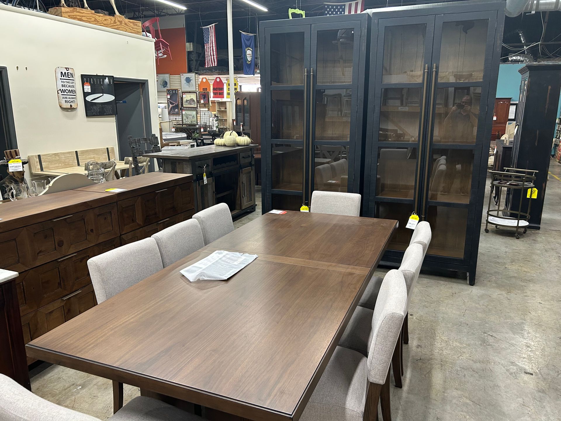 A long wooden dining table with beige chairs in a furniture showroom, with dark display cabinets in the background.