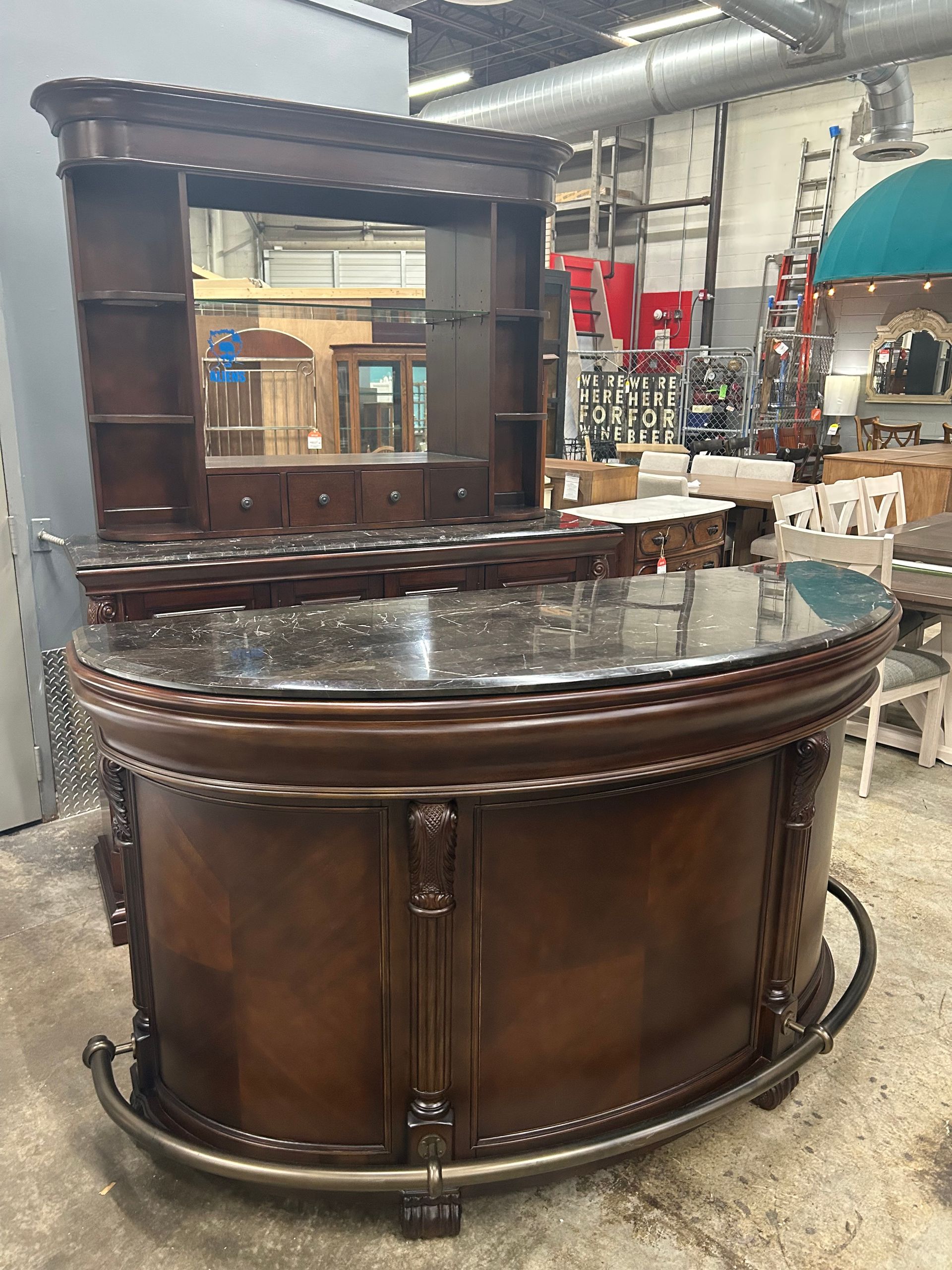 A curved wooden home bar with a dark marble top and a matching overhead hutch, displayed in a showroom setting.