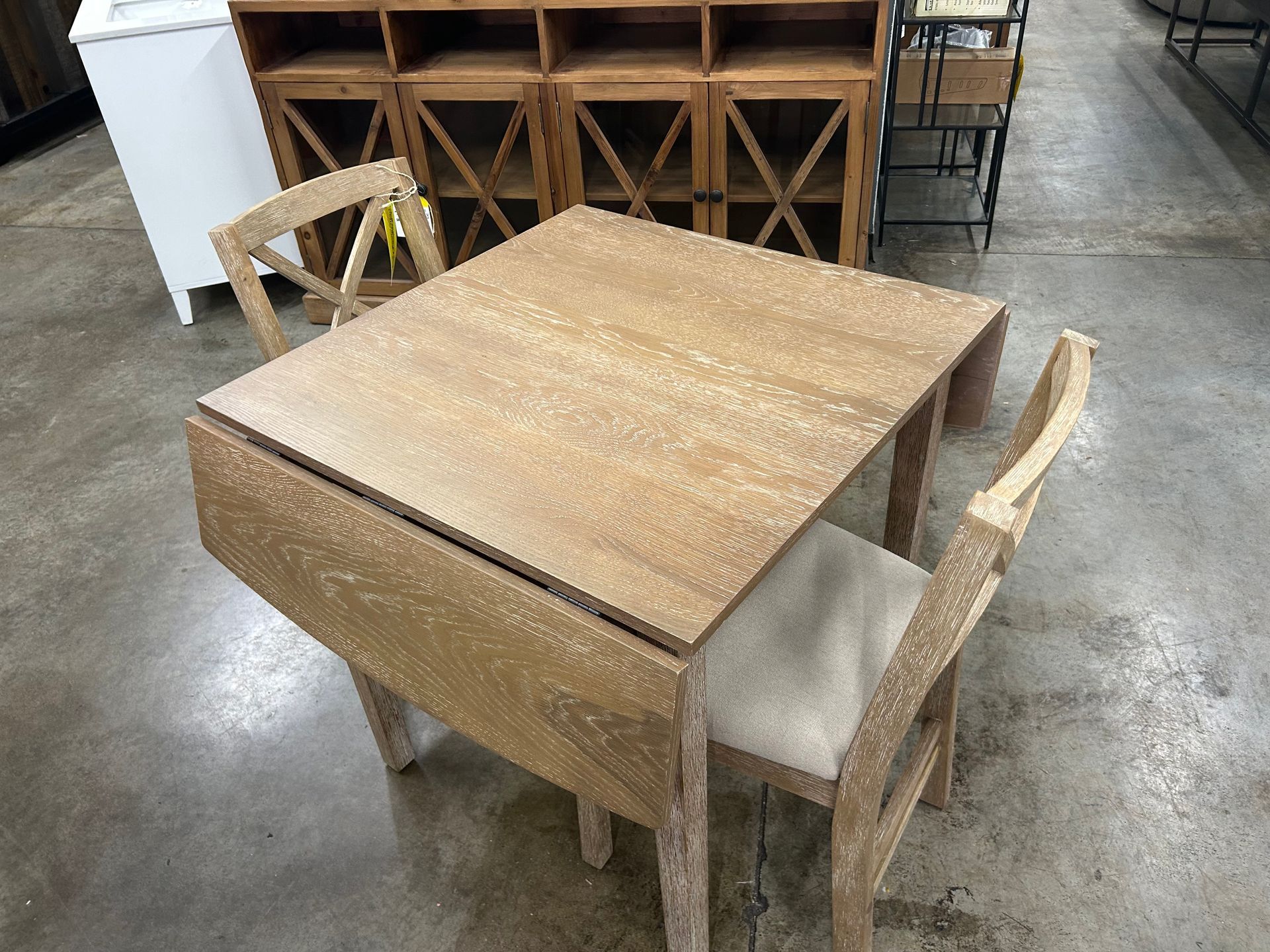 Light wood drop-leaf dining table with two matching chairs, positioned in a retail store setting.