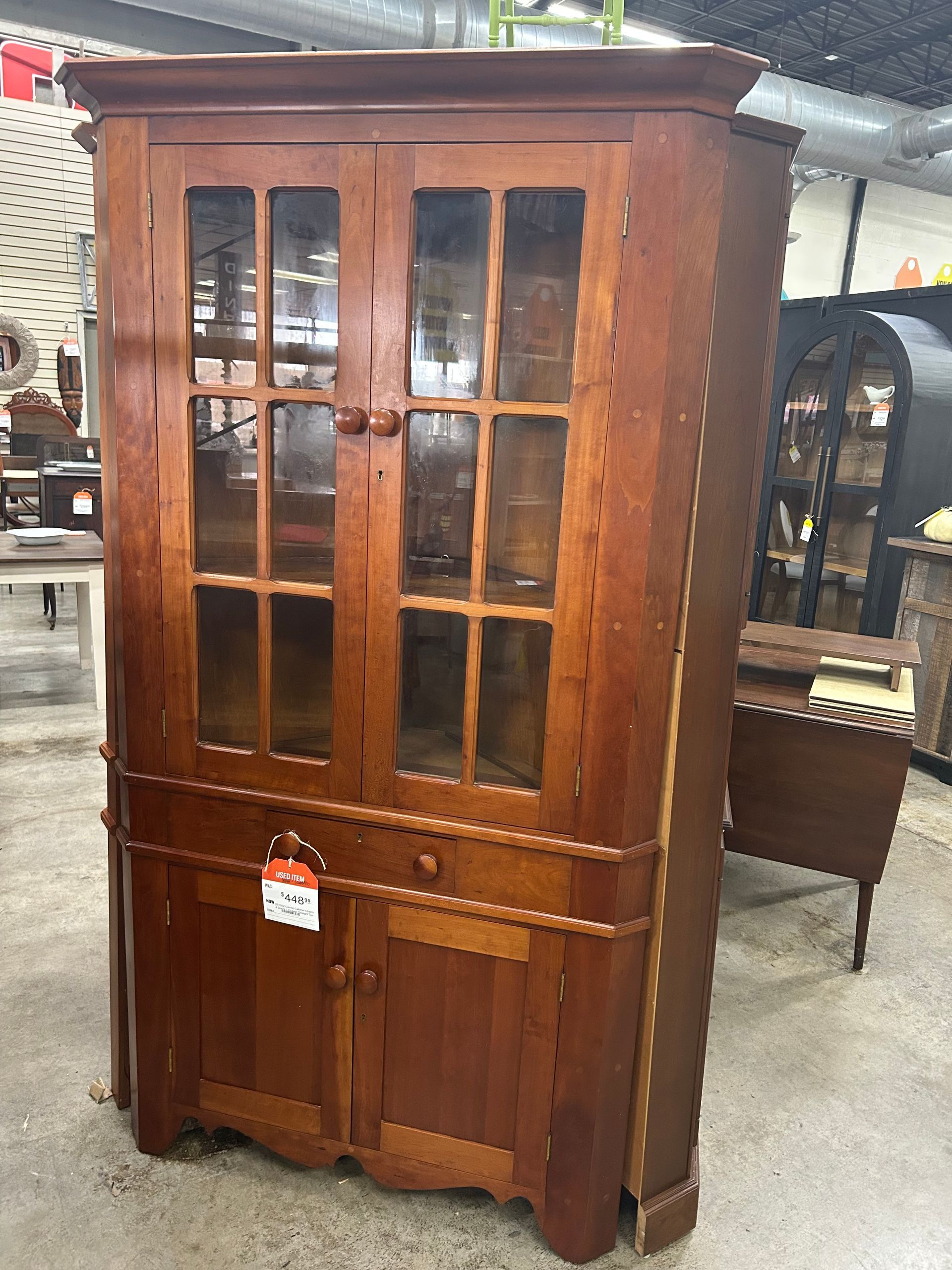 A tall, wooden corner hutch with glass cabinet doors above and a solid wood cabinet base in a retail store setting.