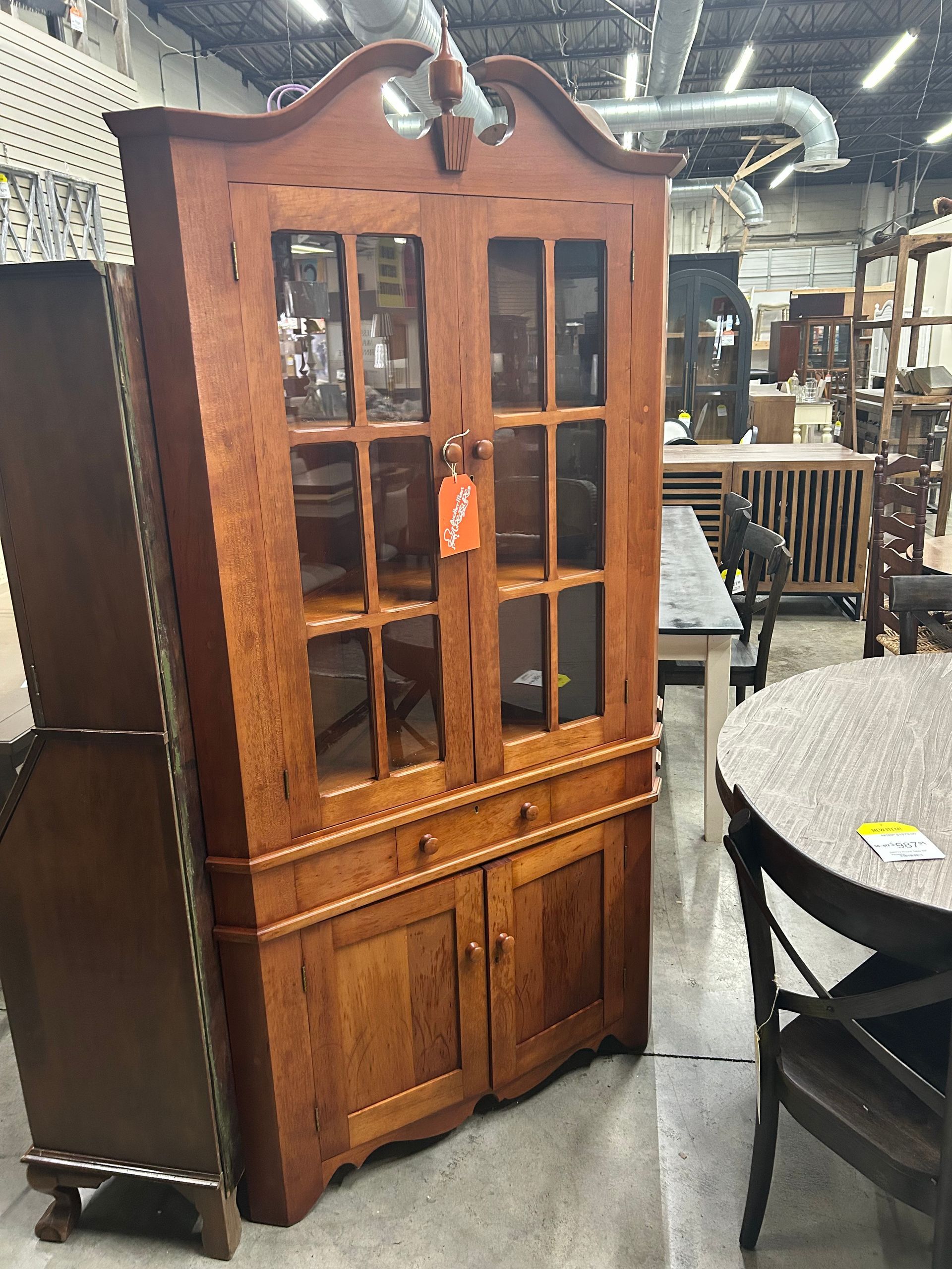 A tall, honey-colored wooden corner hutch with glass-paneled upper doors and lower cabinet storage in a furniture store.