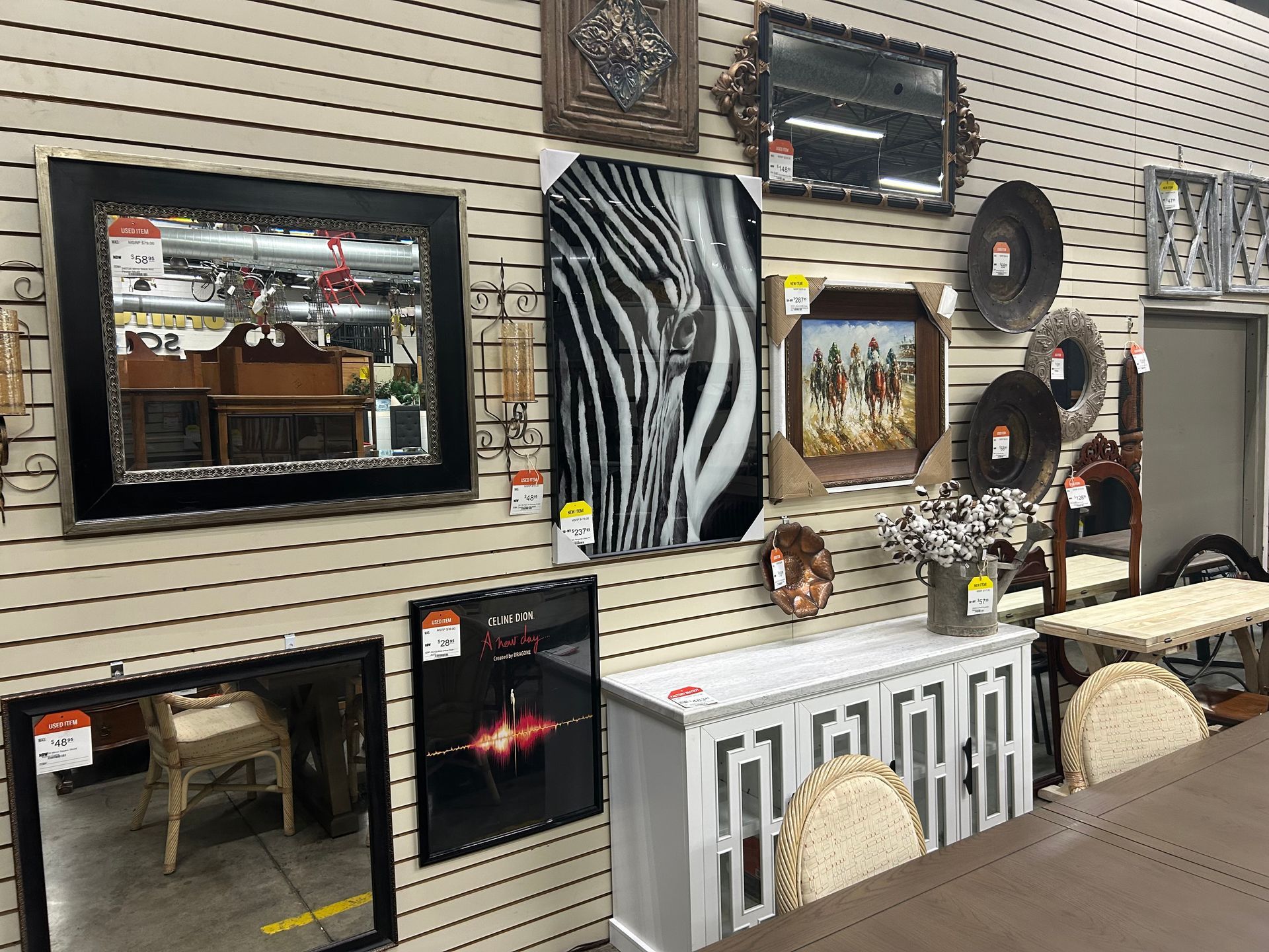 A wall display in a store featuring framed mirrors, artwork, wall decor, and a white cabinet with decorative chairs.