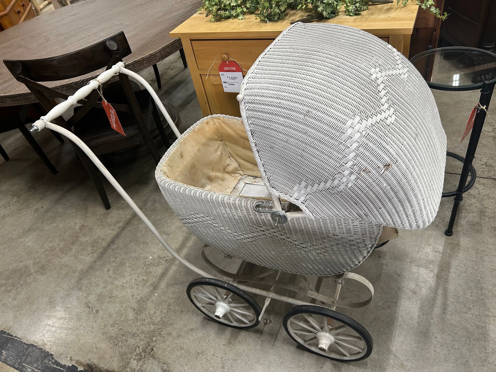 A vintage-style white wicker doll pram with a patterned hood, sitting on a concrete floor in a retail store setting.