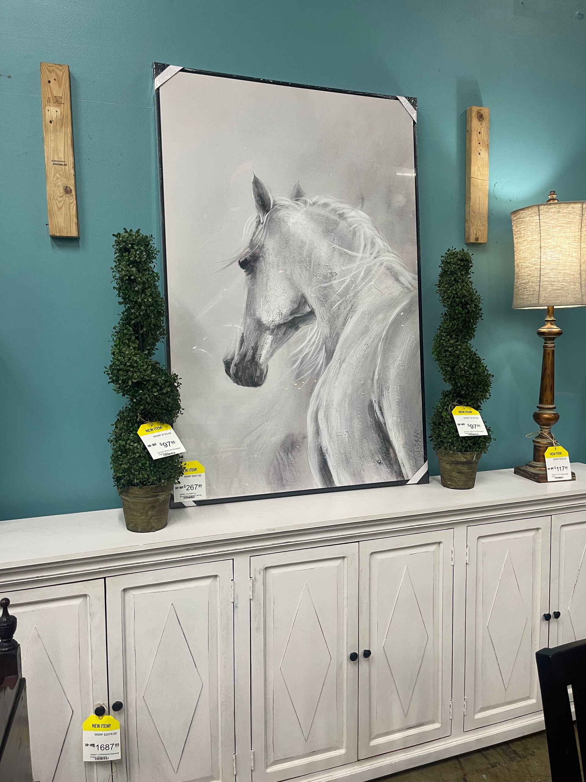 A large painting of a horse centered above a white wooden sideboard, flanked by two spiral topiary plants and a lamp.