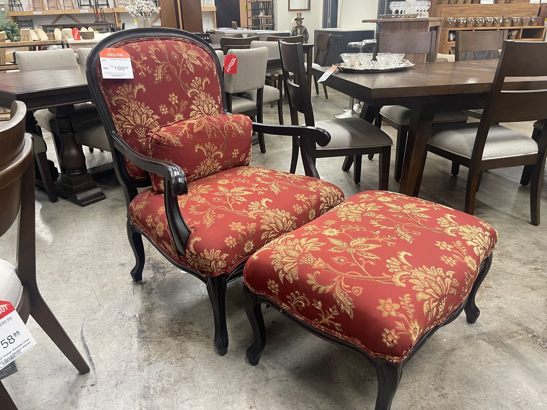A red and gold floral upholstered armchair and matching ottoman with dark wood legs in a furniture showroom.