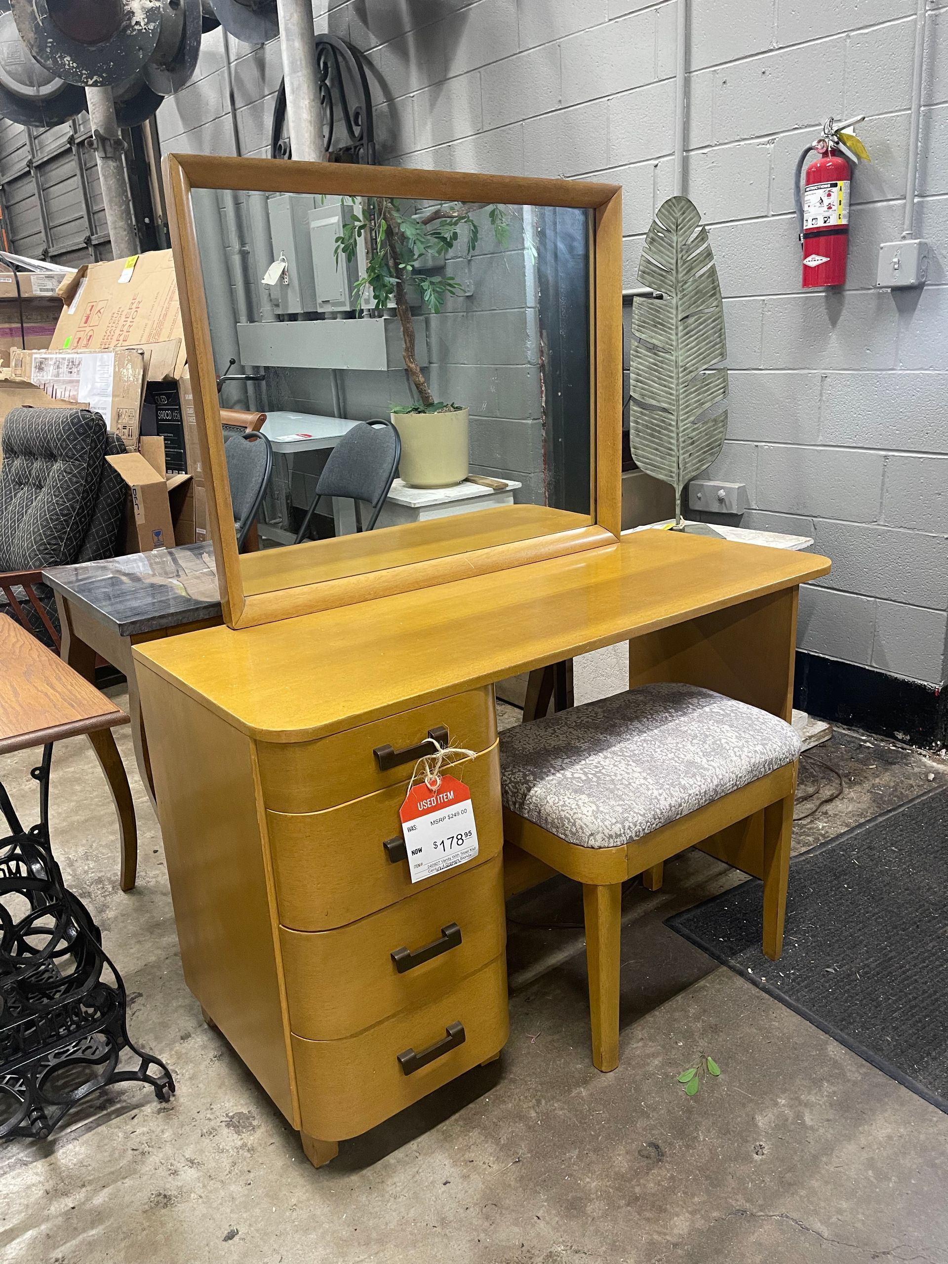 A vintage, light-wood vanity with an attached mirror, three drawers, and a matching fabric-cushioned stool.
