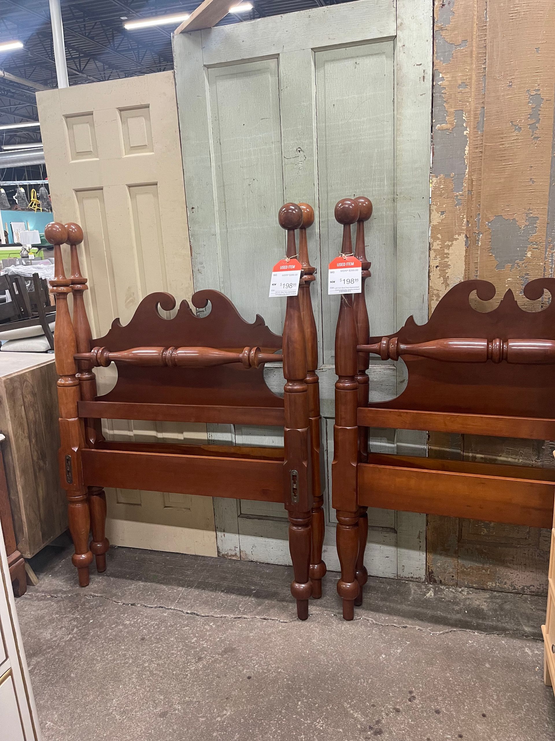 Two matching wooden twin bed headboards with turned posts, standing in a warehouse store with doors in the background.