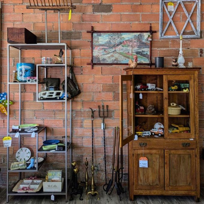 An antique shop display with a metal shelving unit, a wood cabinet, and vintage home goods against a brick wall.
