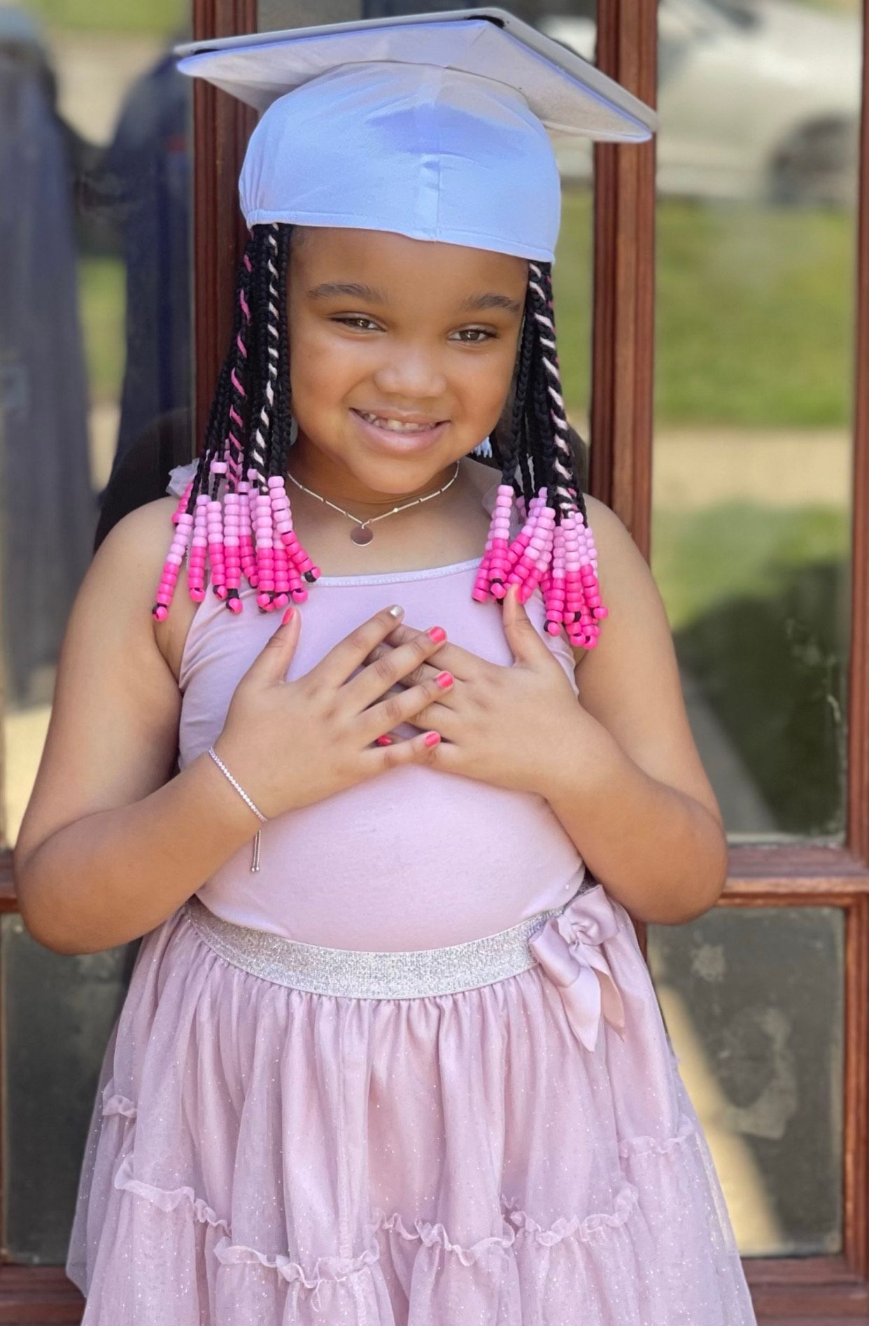 A little girl wearing a graduation cap and gown is standing in front of a window.