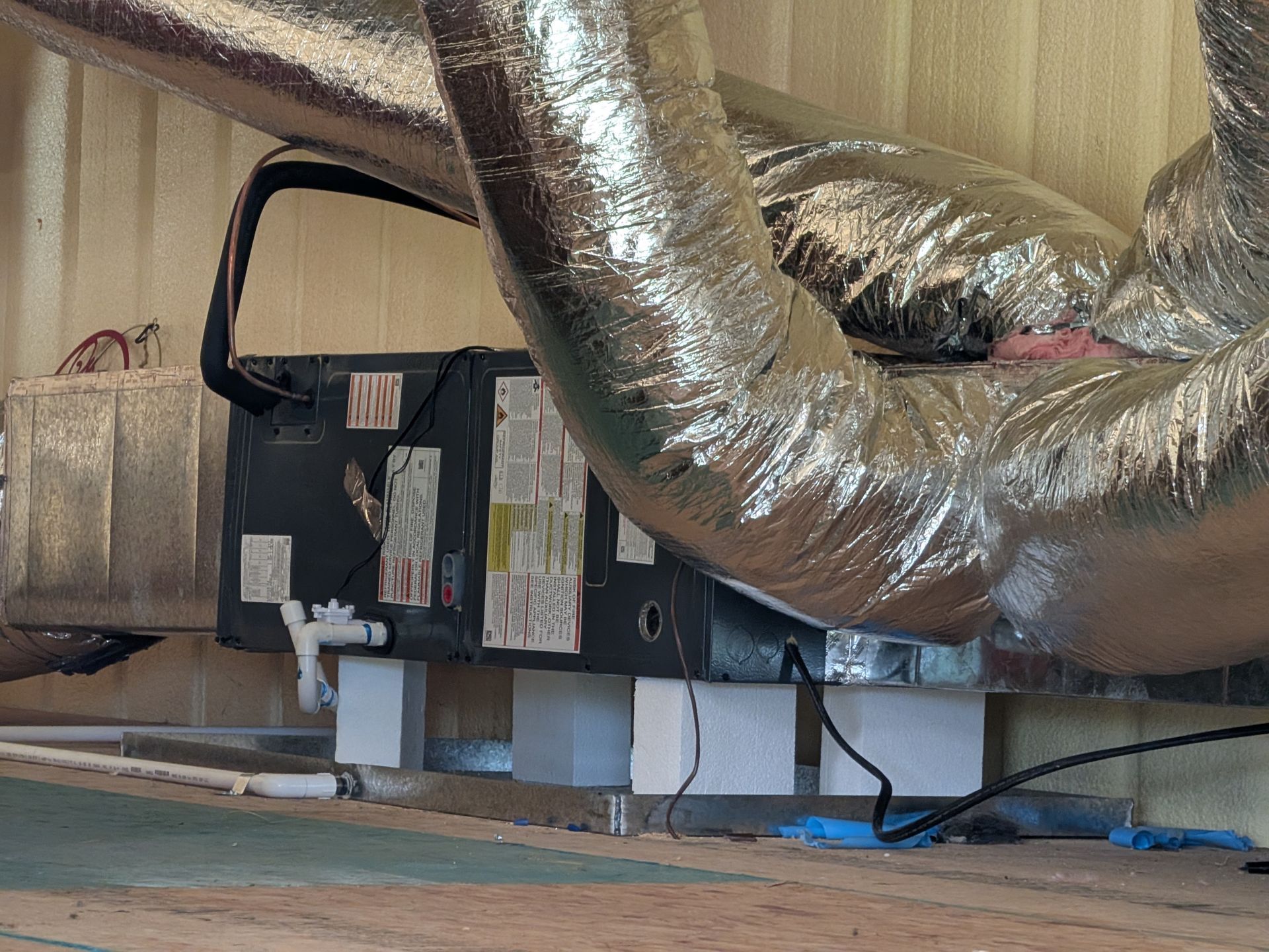 HVAC unit with insulated ducts and white supports against a wall in a room.