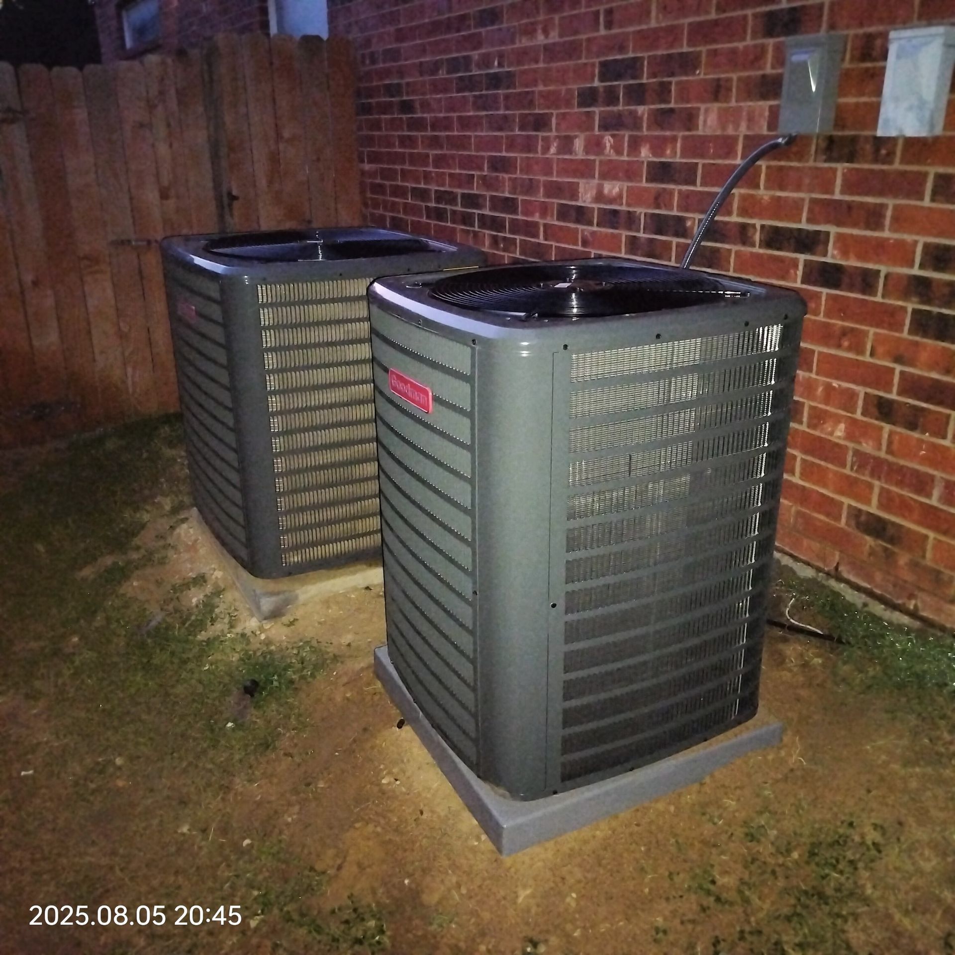 Two outdoor air conditioning units sit on concrete pads next to a brick wall and wooden fence.