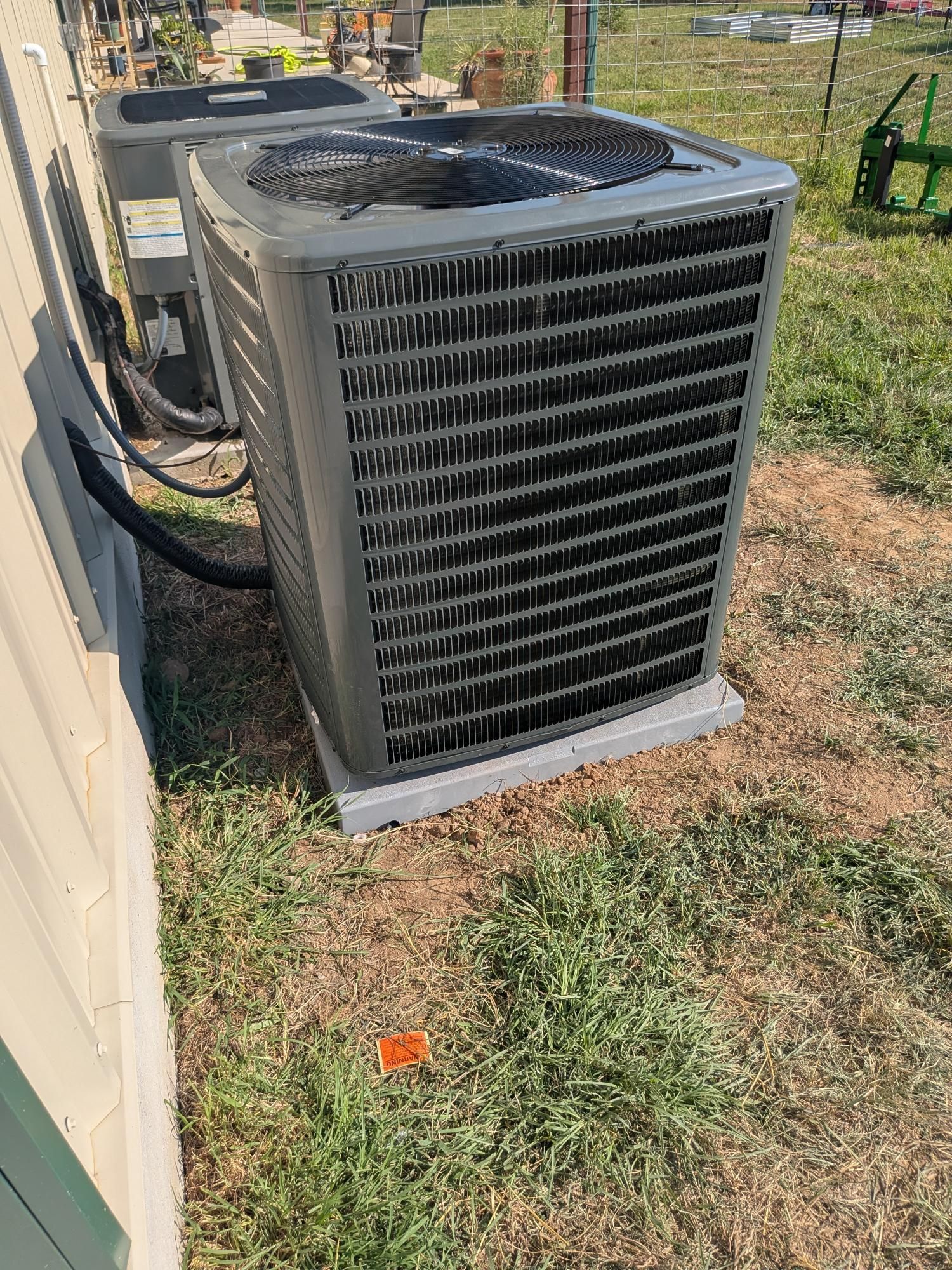 An air conditioning unit on a concrete pad outside a building, surrounded by grass and a fence.