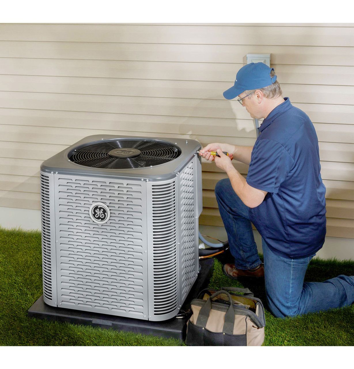 A man in blue kneels by a GE air conditioner on a black pad, using a screwdriver; tool bag sits beside him.