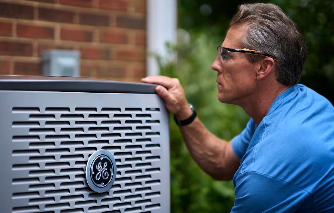 Man in glasses examines a GE air conditioner unit outdoors next to a brick wall.