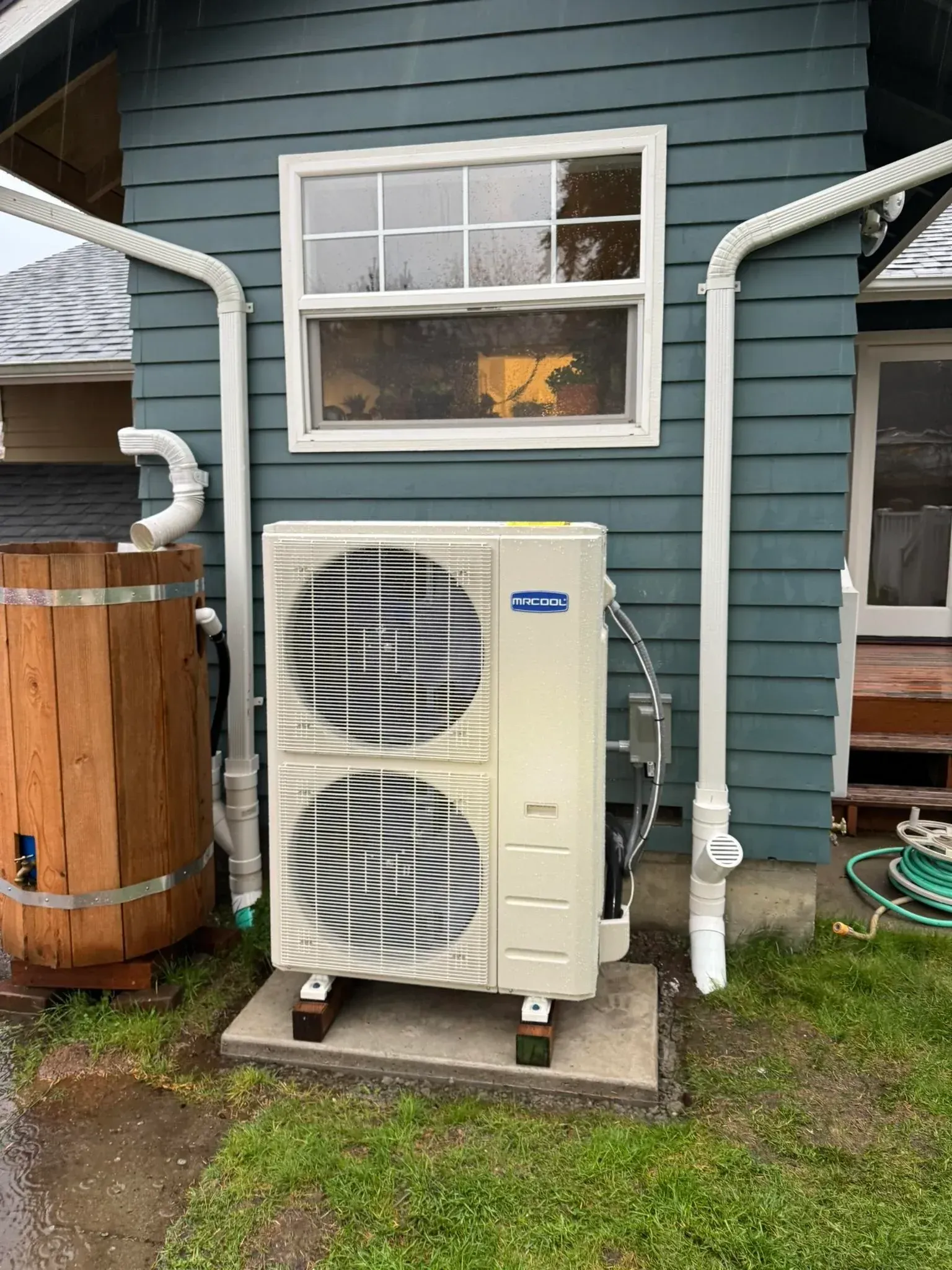 An outdoor heat pump unit against a blue building with white gutters and a small window.