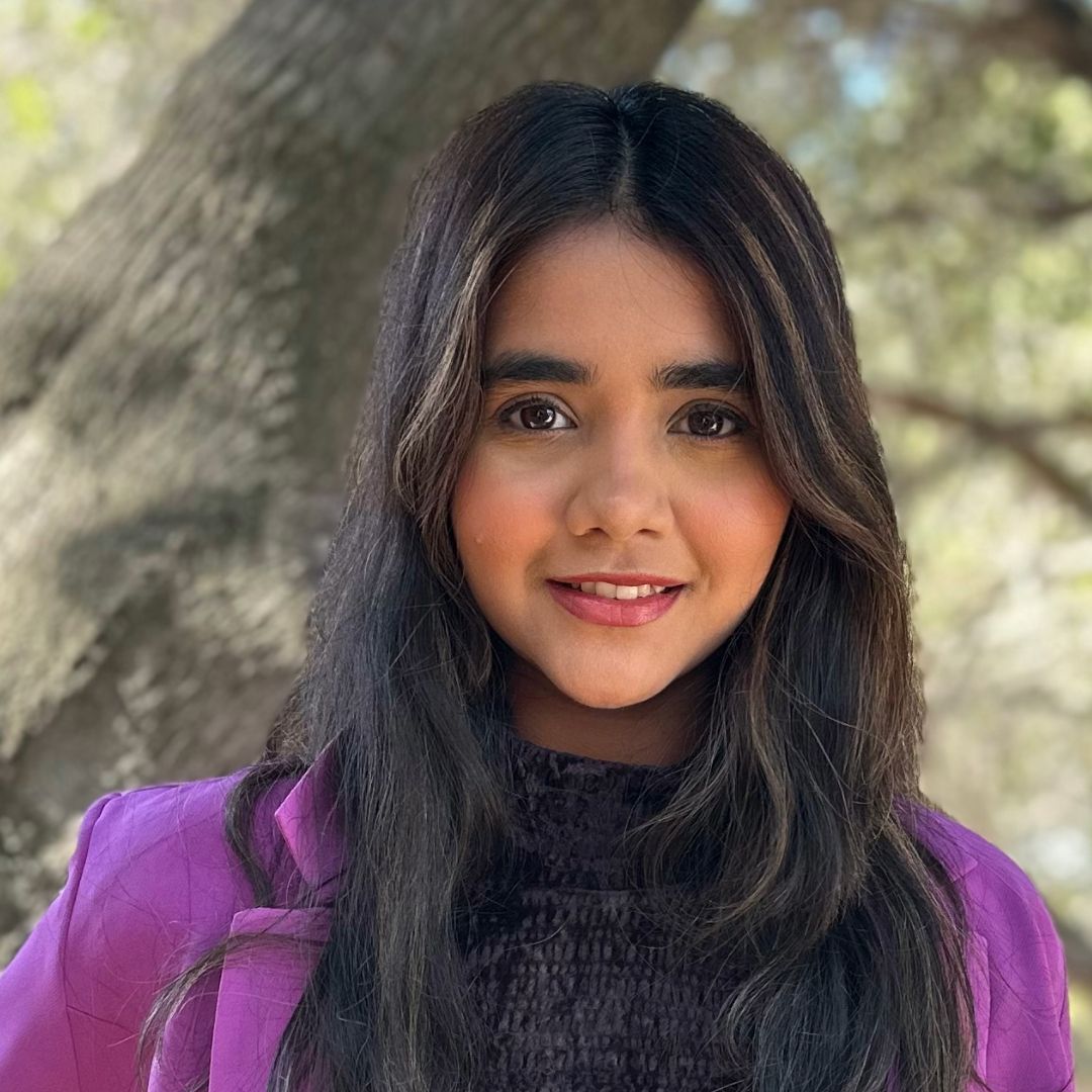 A young woman in a purple jacket is standing in front of a tree.