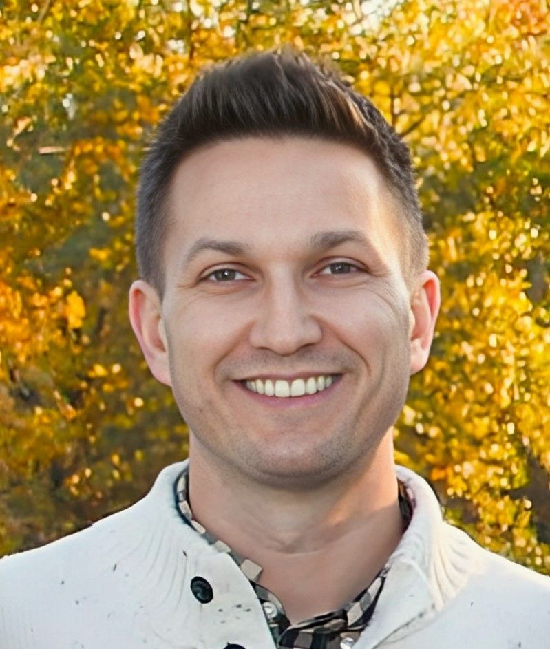 A man wearing a white sweater and a plaid shirt is smiling for the camera.