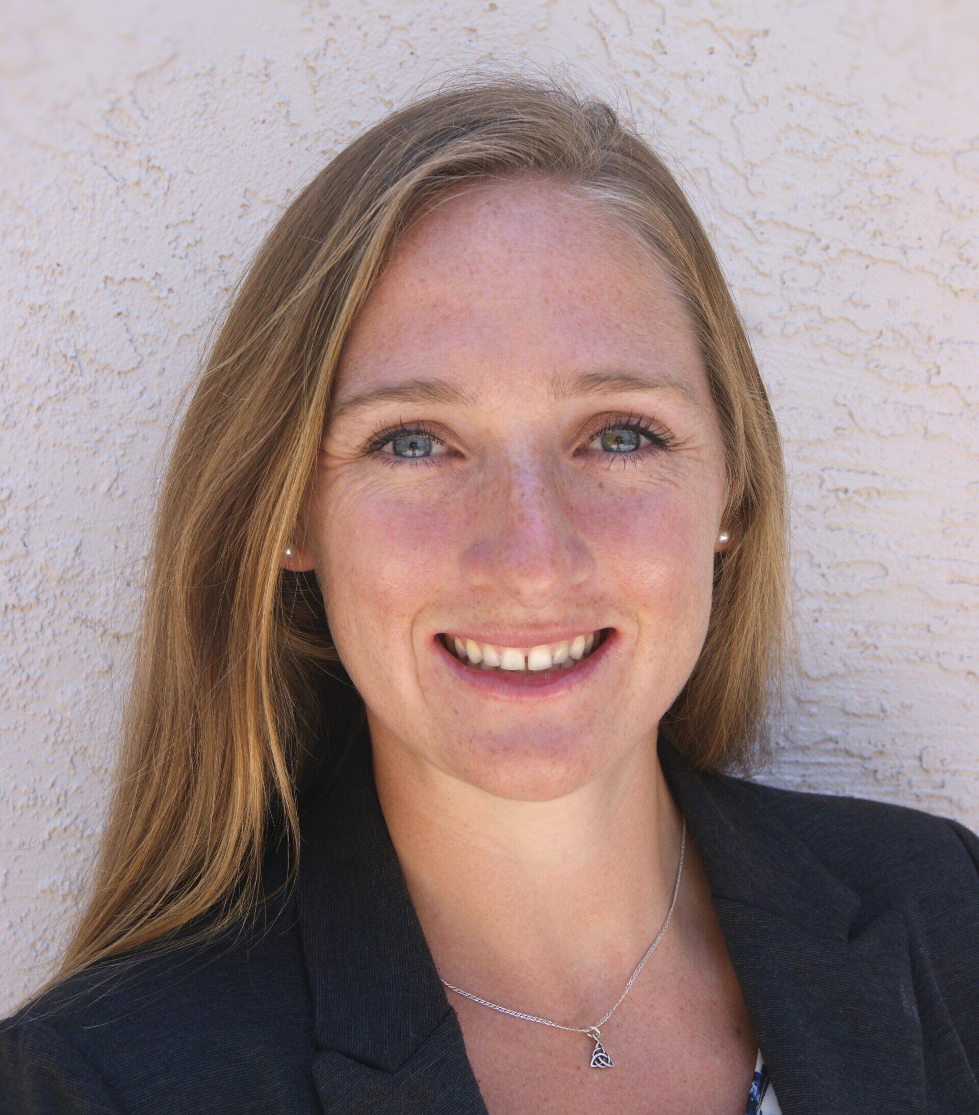 A woman wearing a black jacket and a necklace smiles for the camera