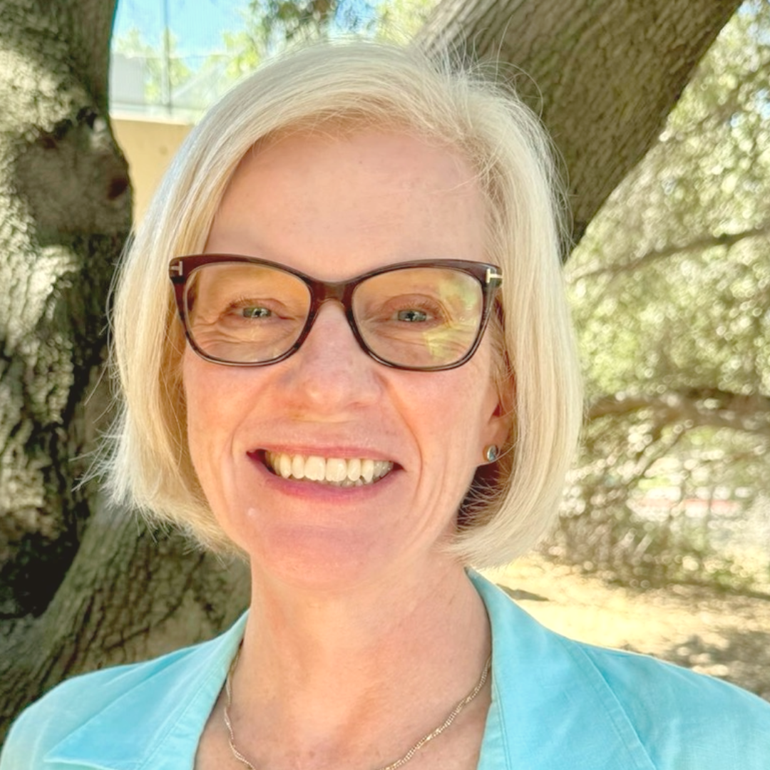 A woman wearing glasses and a blue shirt is smiling in front of a tree.