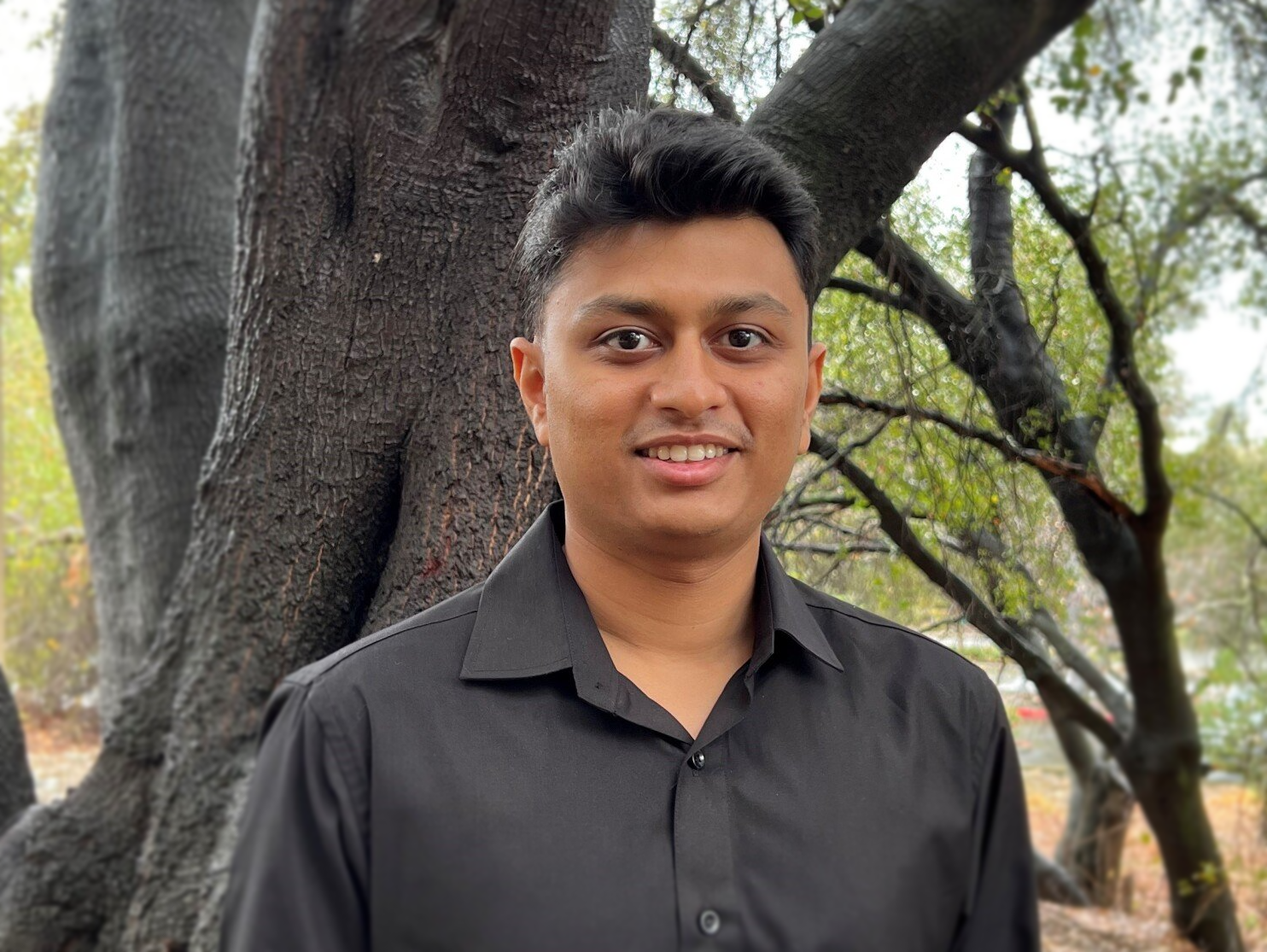 A man in a black shirt is standing in front of a tree.