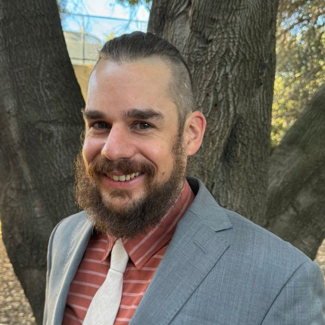 A man with a beard wearing a suit and tie is smiling in front of a tree.