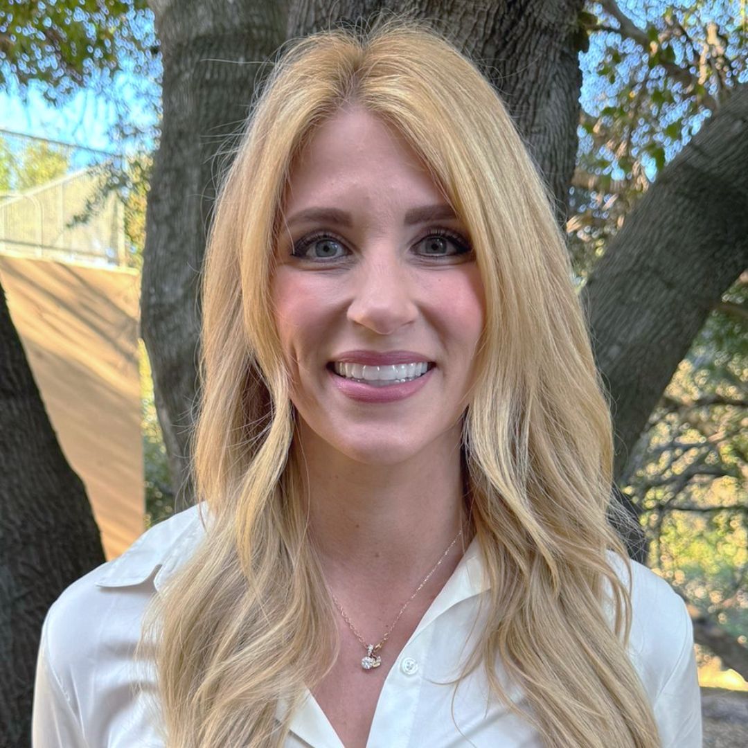 A woman in a white shirt and necklace is smiling in front of a tree.