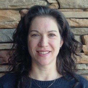 A woman in a blue sweater is smiling in front of a stone wall.