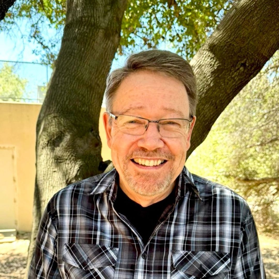 A man wearing glasses and a plaid shirt is smiling in front of a tree.