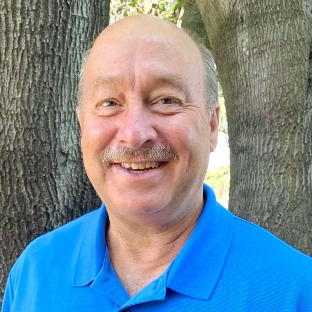 A man with a mustache is wearing a blue shirt and smiling in front of a tree.