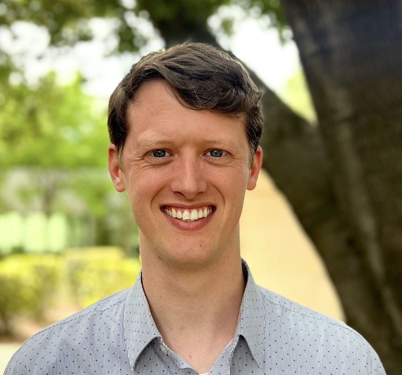 A man in a striped shirt is smiling in front of a tree