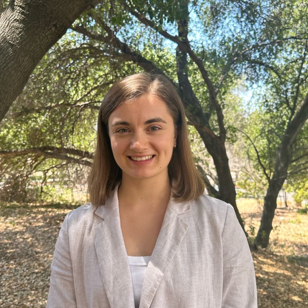 A woman in a white jacket is smiling in front of trees.