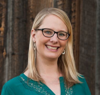 A woman wearing glasses and earrings is smiling in front of a wooden wall.