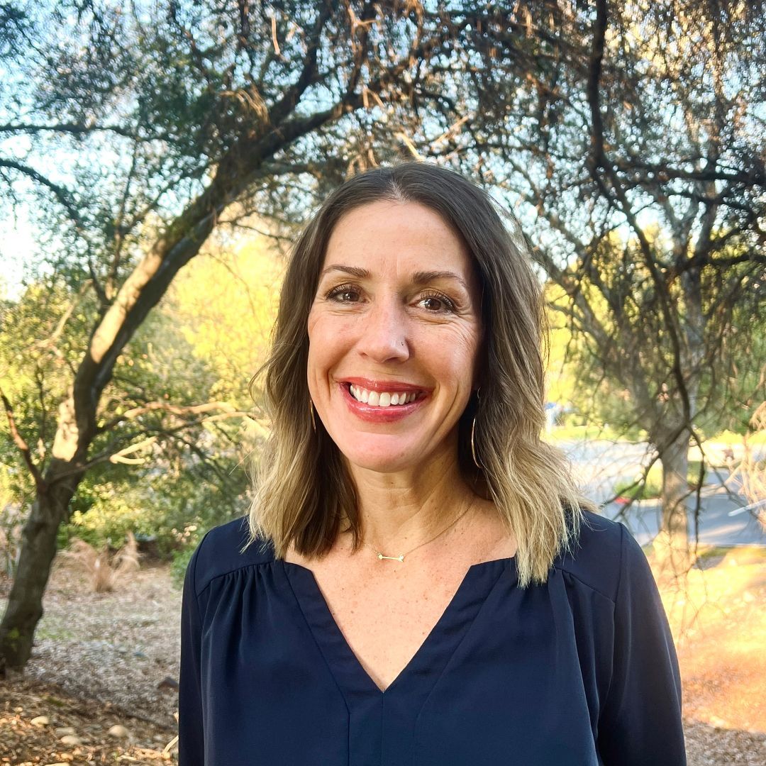 A woman in a suit is smiling in front of a tree.