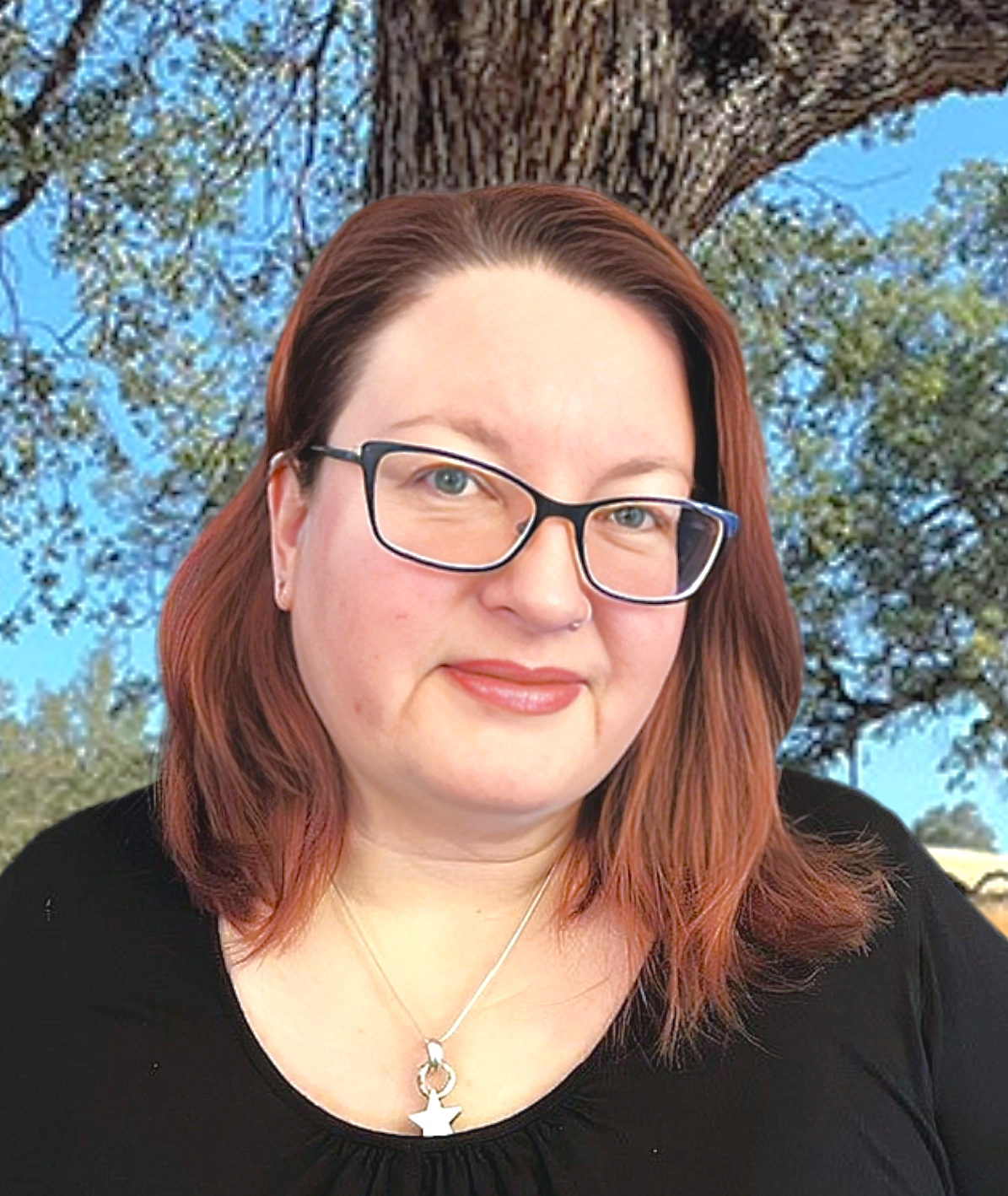 A woman wearing glasses and a necklace is standing in front of a tree.