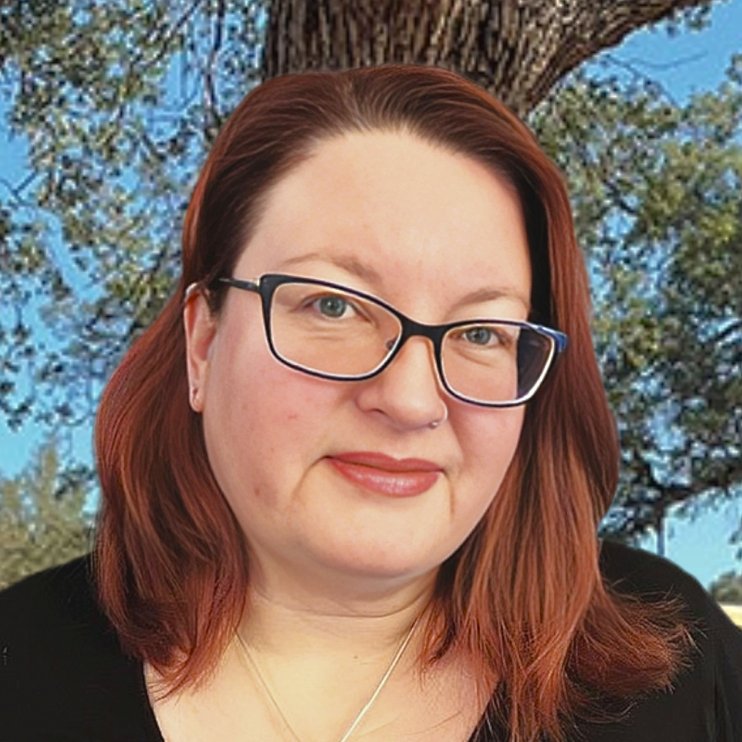 A woman with red hair and glasses is standing in front of a tree.