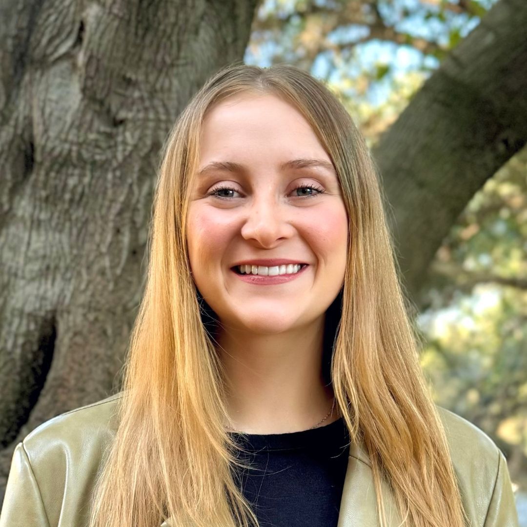 A woman with long blonde hair is smiling in front of a tree.