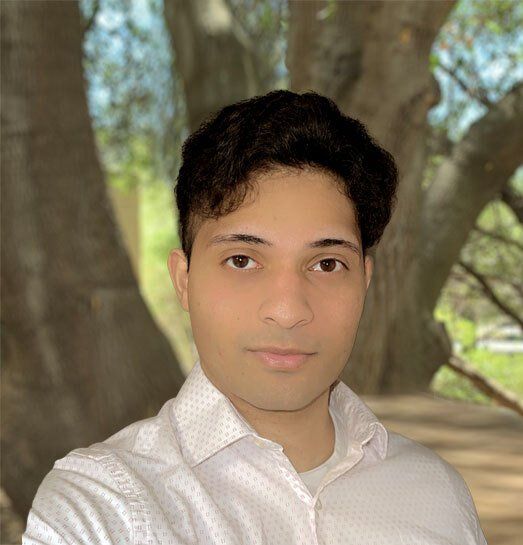 A young man in a white shirt is standing in front of a tree.