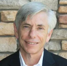 A man in a suit and white shirt is smiling in front of a brick wall.