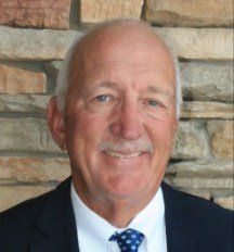 A man in a suit and tie is smiling in front of a stone wall.