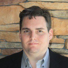 A man in a suit and blue shirt is standing in front of a stone wall.