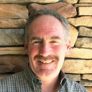 A man with a mustache is smiling in front of a stone wall.