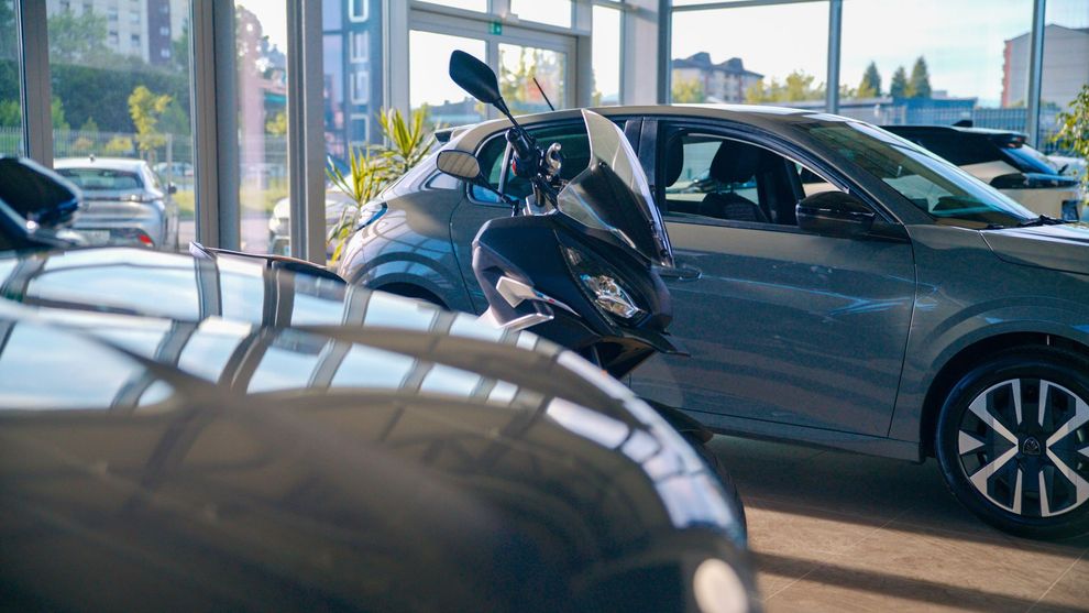 Auto e una motocicletta esposte all'interno dello showroom di una concessionaria con luce naturale.