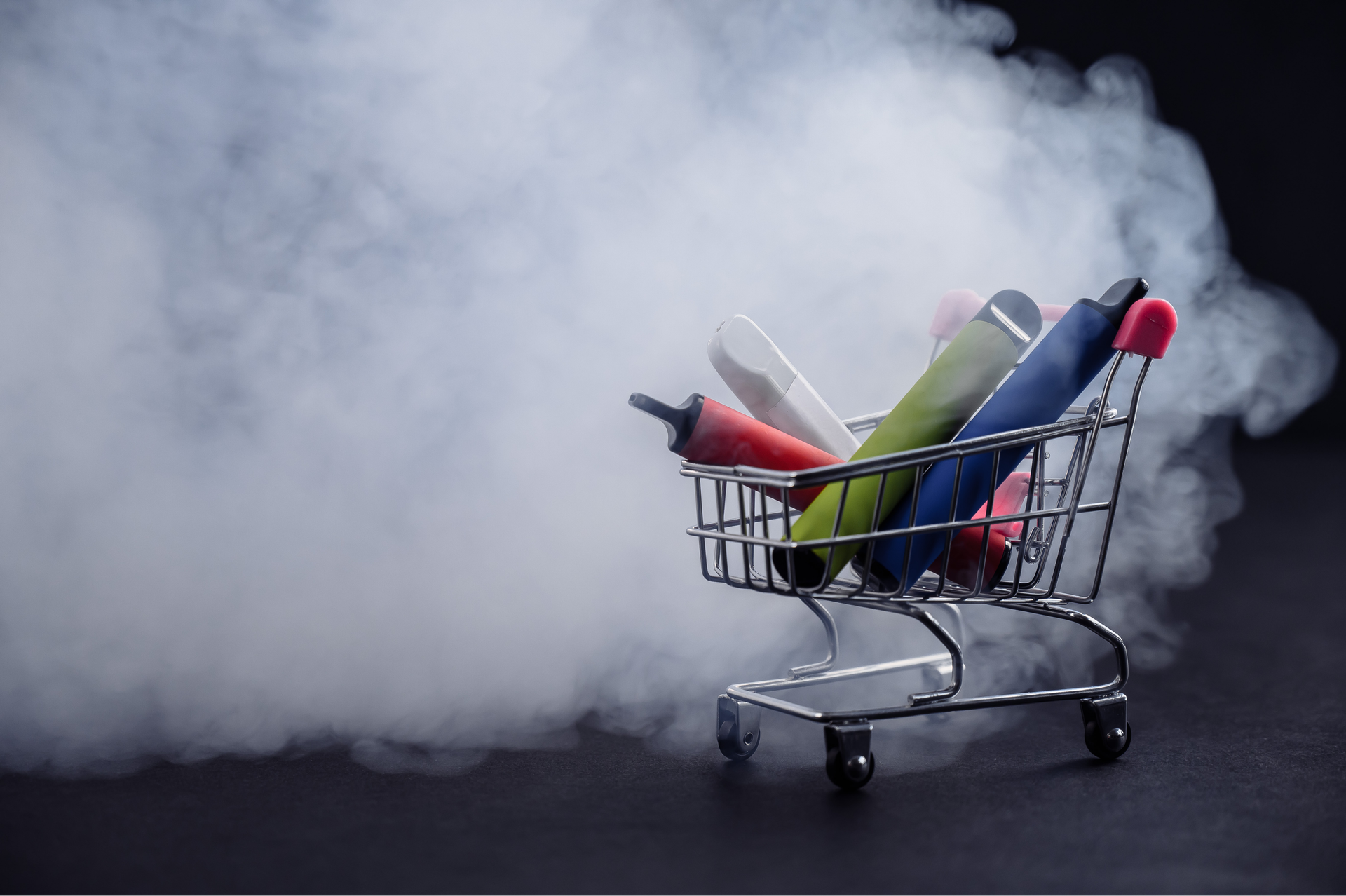 Mini shopping cart filled with colorful disposable vapes, surrounded by white vapor.