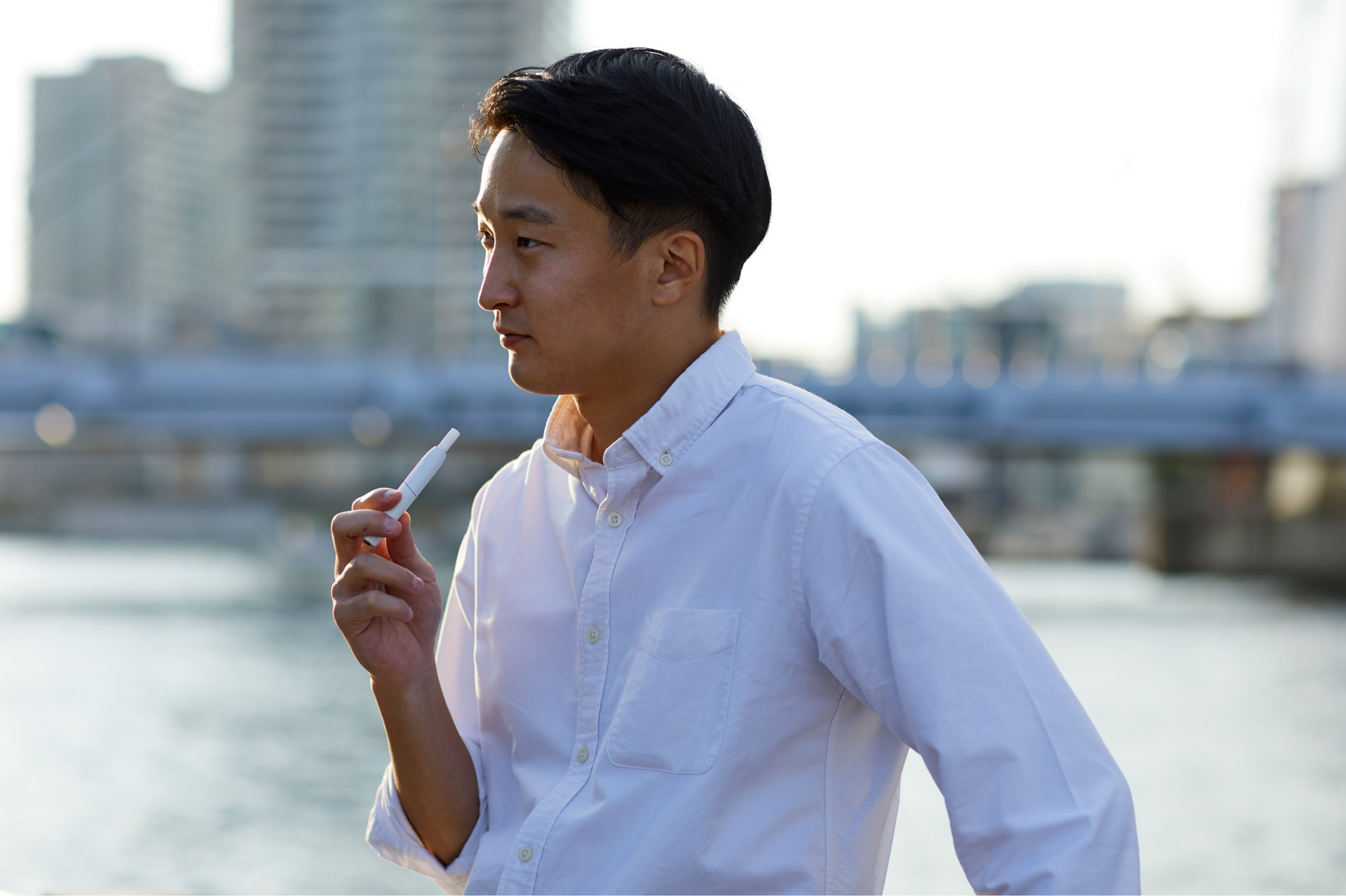 Man in white shirt holds a white object, standing outdoors by water with a city backdrop.