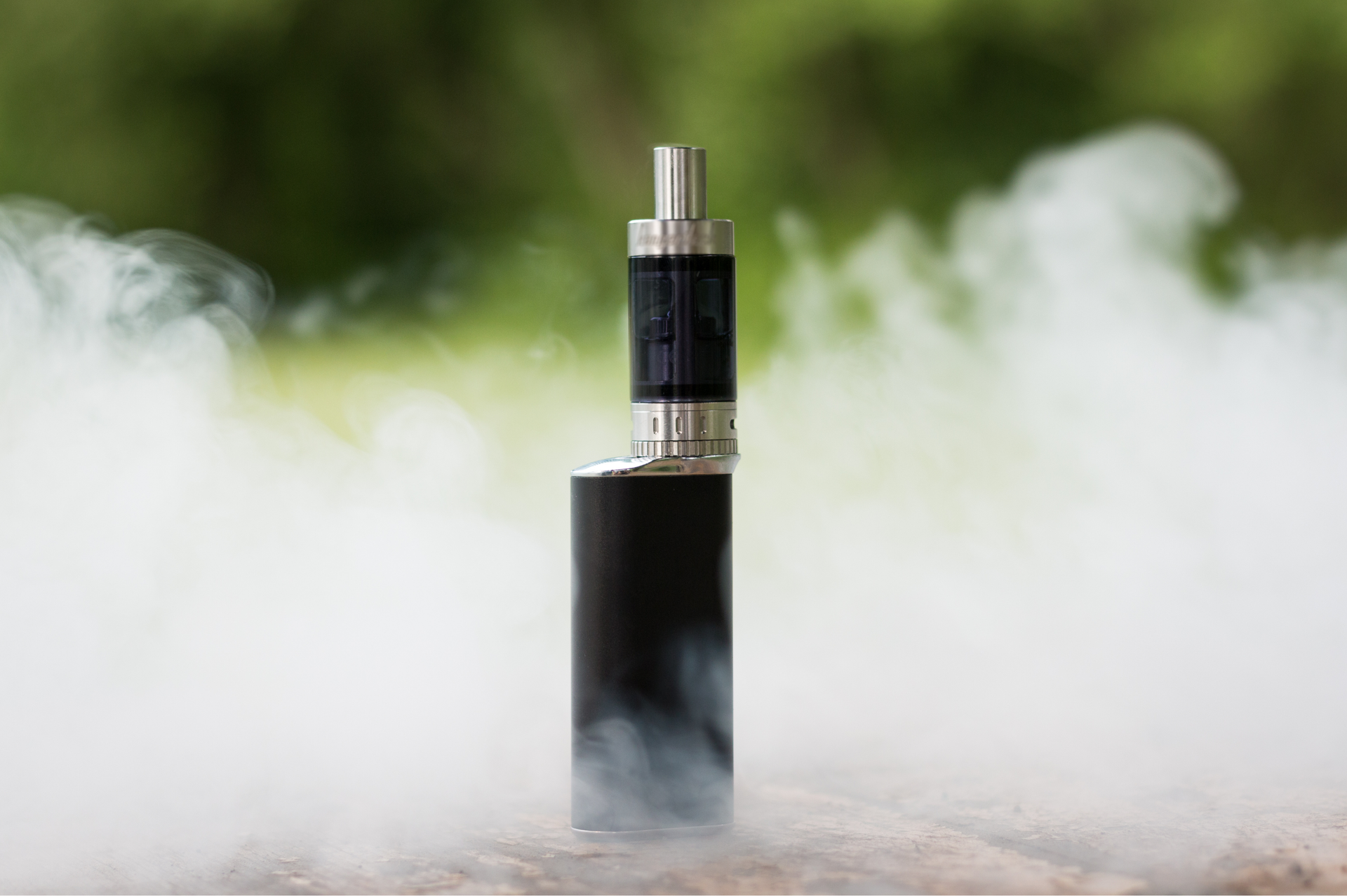 Black vape device emitting a cloud of vapor outdoors with blurred green background.