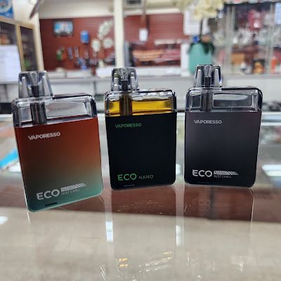 Three Vaporesso Eco Nano vape devices: orange, green, and black, on a glass surface in a store.