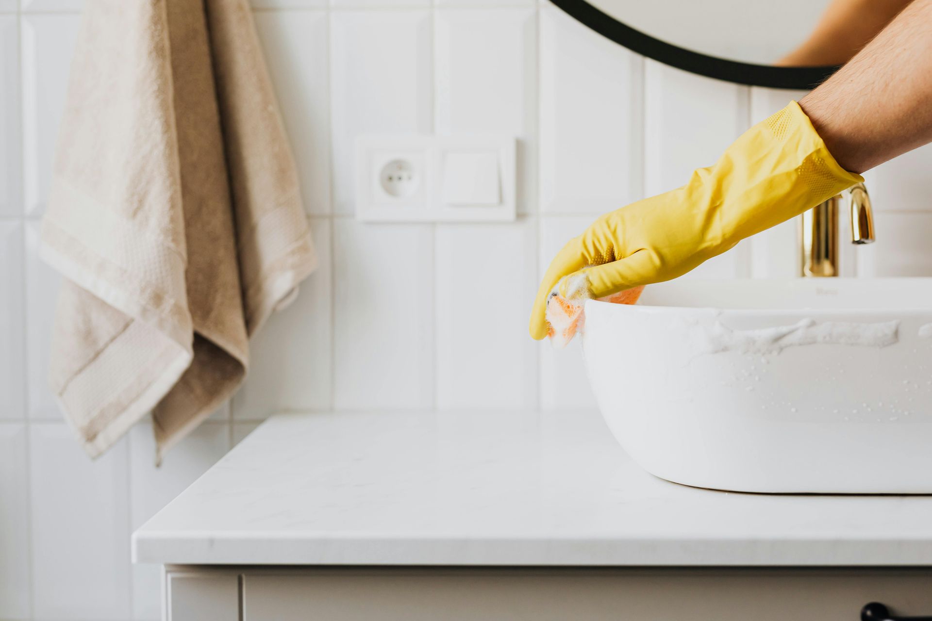 A person wearing yellow gloves is cleaning a bathroom sink with a sponge.