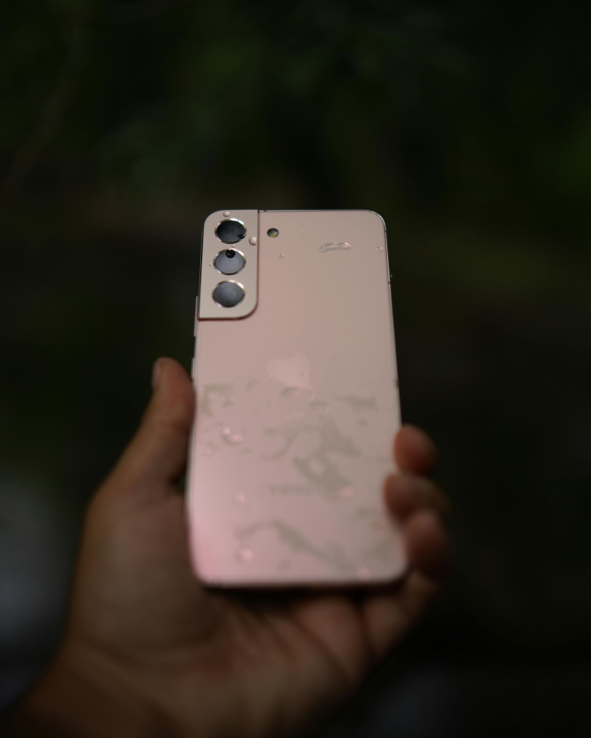 Eine Hand hält ein pinkfarbenes Samsung-Smartphone mit Wassertropfen. Drei Kameras oben, dunkler Hintergrund.